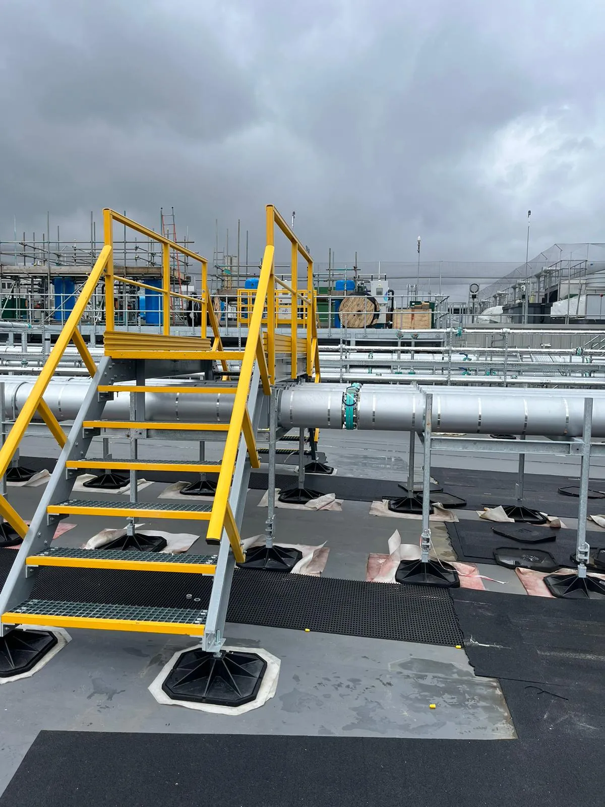 Roof Access, Stepovers & Edge Protection Systems
Safe pedestrian access at roof level is essential for ongoing inspection and maintenance, particularly where mechanical and electrical equipment is housed in rooftop service areas.
We supply walkway systems that provide safe, clearly defined pedestrian routes across roof surfaces to plant rooms. These clearly defined walkways help prevent accidental slips and falls while protecting roof membranes and finishes.
GRP walkways are especially well suited to roof environments due to their slip-resistant surface, corrosion resistance and low maintenance requirements, offering a durable solution for long-term access. GRP or steel handrails provide continuous walkway edge protection.
A range of lightweight, easy to install GRP rooftop stepovers are readily available. These step-over platforms are essential to provide safe access over obstructions and hazards on a roof, such as pipes, cable trays, HVAC ductwork, or changes in roof level. They enhance worker safety by providing a secure, stable, and non-slip walkway that prevents trip hazards and falls.
Our roof edge protection systems provide fall protection along roof perimeters, helping prevent falls from height in accordance with industry regulations. Available as freestanding, non-penetrative systems or fixed installations, these modular steel systems can be configured to suit a wide range of roof types and finishes.
Together, handrailed walkways and edge protection systems can be combined to create safe, roof access solutions, supporting the movement of personnel from access points to M&E service rooms and plant.